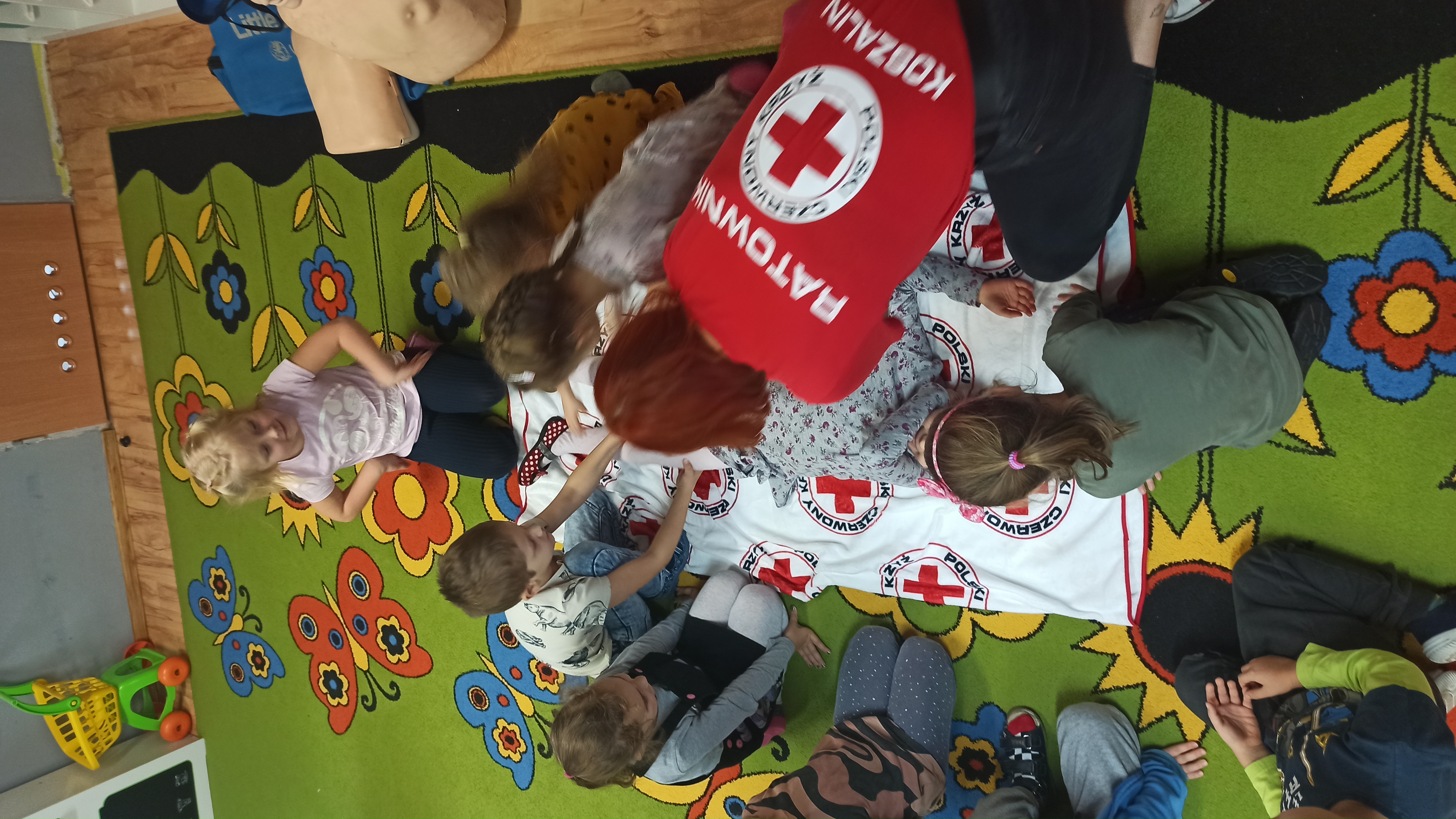 Zdjęcie przedstawia przedszkolaki uczestniczące w zajęciach z pierwszej pomocy wraz  z ratownikami. 